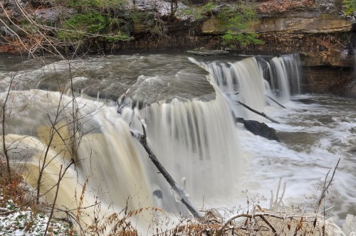 A serene waterfall cascades over rocky ledges, surrounded by trees and a tranquil river.