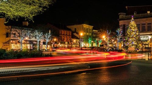 A festive town square at night, featuring holiday lights, a decorated tree, and light trails from passing cars.
