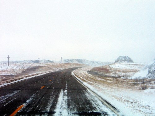 A snowy, winding road stretches through a desolate landscape with hills in the distance and a gray sky.