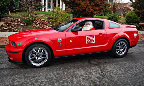 A red sports car with Santa inside, decorated with colorful stickers, parked in a festive outdoor setting.
