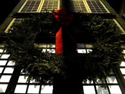 A large green wreath with a red bow hangs on a building, illuminated against a dark night sky.
