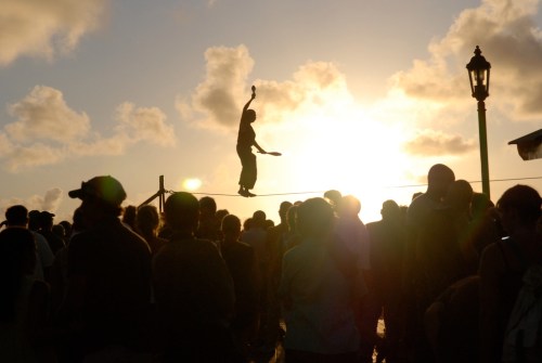 A performer balances on a tightrope against a sunset, surrounded by a crowd of onlookers.