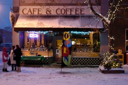 A cozy café with a "OPEN" sign, surrounded by snow, with people chatting outside in winter attire.