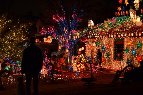 A person stands in front of a house adorned with colorful Christmas lights and decorations, creating a festive atmosphere.