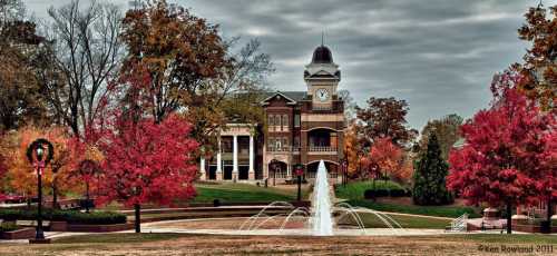 A fountain in a park surrounded by vibrant autumn trees and a historic building with a clock tower in the background.