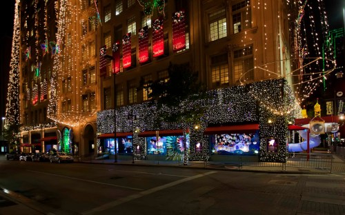 A festive city street at night, adorned with colorful holiday lights and decorations on buildings and storefronts.