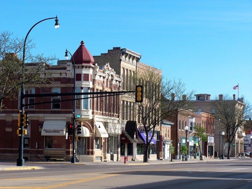 Historic buildings line a quiet street under a clear blue sky, with traffic lights and trees in the foreground.