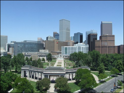 A panoramic view of a city skyline with modern buildings and a green park in the foreground. Clear blue sky above.