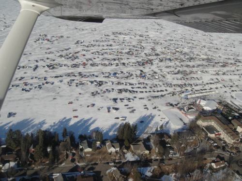 Aerial view of a snowy landscape filled with parked cars, with a small town visible at the bottom of the image.