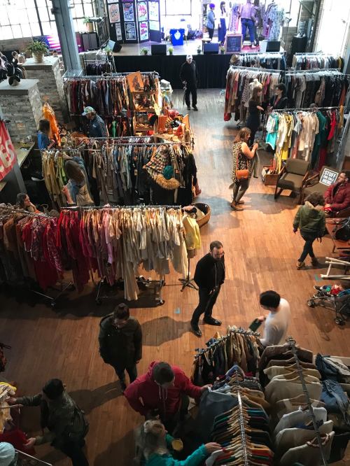 A bustling vintage market with racks of clothing, shoppers browsing, and a stage in the background.