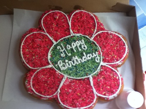 A large flower-shaped birthday cake decorated with colorful sprinkles and the words "Happy Birthday" in the center.
