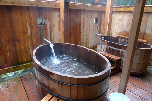 Two wooden soaking tubs filled with water, surrounded by wooden walls and a deck, creating a serene outdoor spa setting.