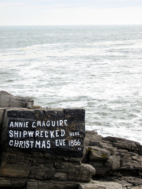 A rocky shoreline with a sign reading "Annie C. Maguire Shipwrecked Here Christmas Eve 1886" against a backdrop of the ocean.