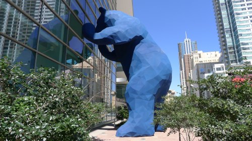 A large blue bear sculpture leans against a glass building, surrounded by greenery and city skyscrapers in the background.