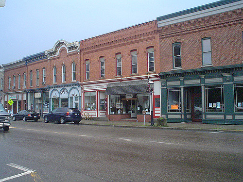 A row of brick storefronts along a street, featuring various shops and parked cars under a cloudy sky.