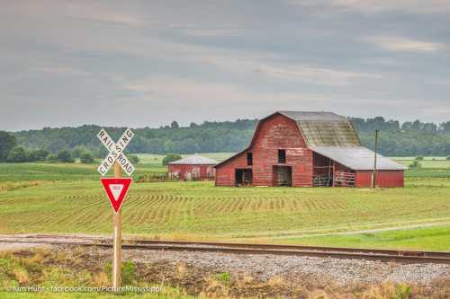 A red barn stands near a railroad crossing in a rural landscape with fields and trees in the background.