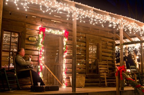 A cozy log cabin decorated with holiday lights, featuring a person sitting on the porch in a rocking chair.