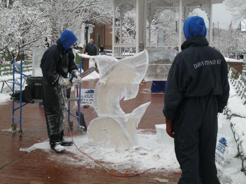 Two artists sculpt ice in a snowy park, one using a chainsaw, while the other observes. Snow-covered trees in the background.