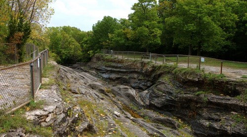 A rocky, layered landscape with a fenced area and trees in the background, showcasing natural erosion and greenery.
