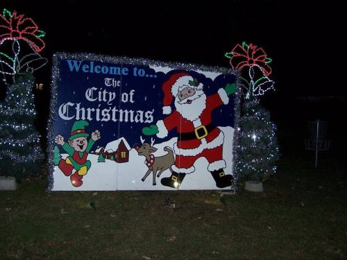 A festive sign reading "Welcome to the City of Christmas," featuring Santa, an elf, a reindeer, and decorated trees.