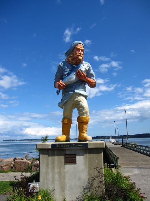 A colorful statue of a fisherman holding a fish, located in Eastport, Maine, with a scenic waterfront in the background.
