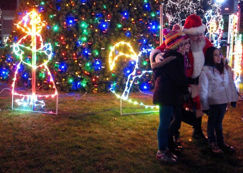 A man in a Santa suit poses with two children in front of colorful holiday lights and decorations.