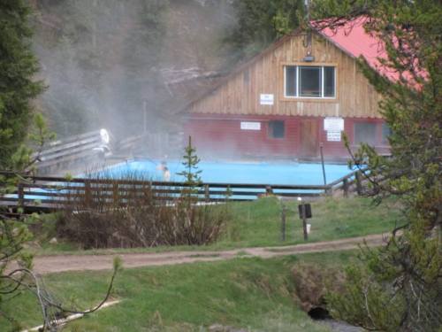 A wooden building near a steaming blue hot spring, surrounded by greenery and a dirt path.