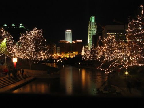 City skyline at night, illuminated buildings, and trees adorned with festive lights reflecting in a calm waterway.