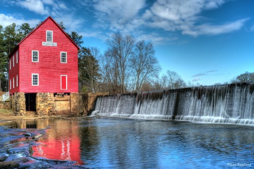 A red historic mill beside a waterfall, reflecting in a calm river under a blue sky with scattered clouds.