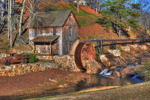 A rustic wooden mill with a large water wheel beside a stream, surrounded by trees and a vintage truck nearby.