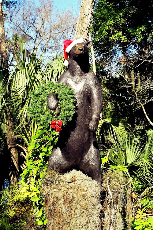 A bear statue wearing a Santa hat holds a Christmas wreath, surrounded by lush greenery.