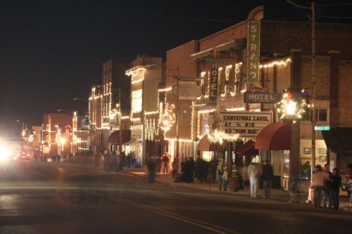 A festive street scene at night, with buildings adorned in holiday lights and people gathered along the sidewalk.