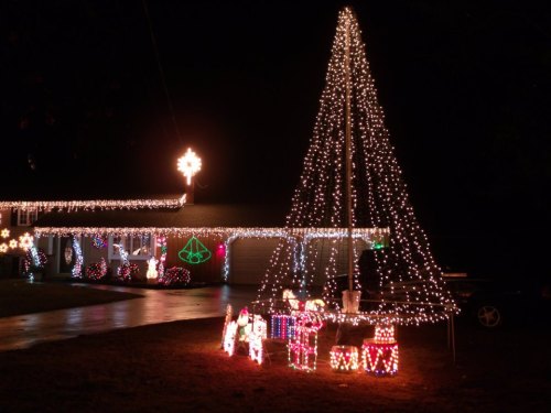 A house decorated with colorful Christmas lights, featuring a large lit tree and festive presents in the yard.