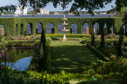 A lush garden with a fountain, surrounded by greenery and arches, overlooking a calm body of water.