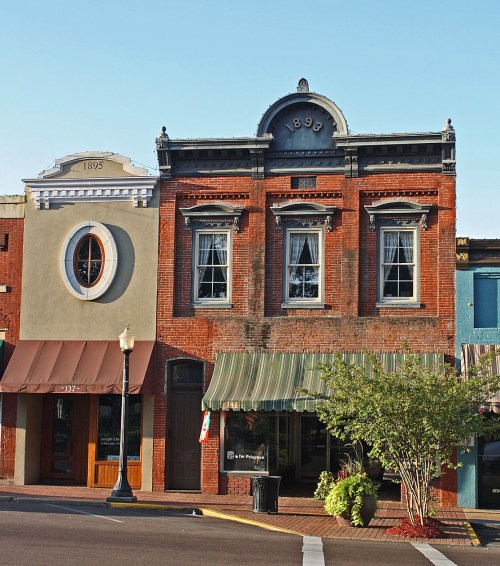 Historic brick buildings with large windows and awnings, featuring a round window and a decorative 1895 sign.