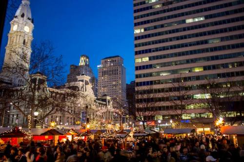 A bustling holiday market at dusk, with festive lights and a historic building in the background.