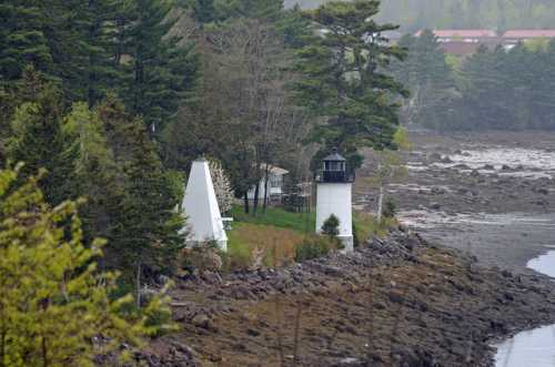 A coastal scene featuring a white lighthouse and a triangular building surrounded by trees and rocky shoreline.