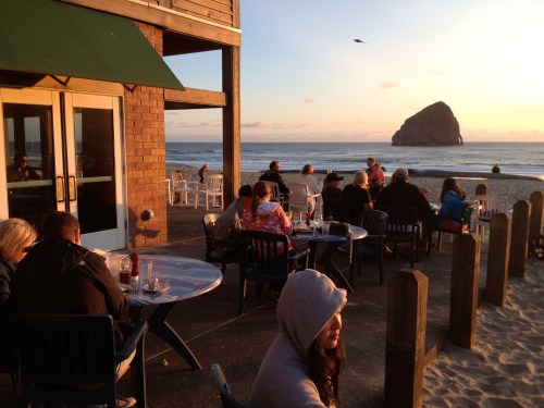 People dining outdoors at a beachside restaurant during sunset, with a large rock formation in the background.