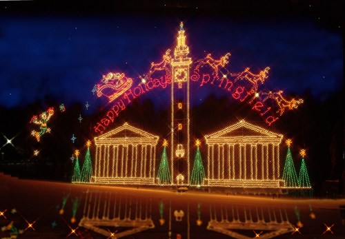 Colorful holiday lights display featuring a tower, trees, and the text "Happy Holidays Springfield." Nighttime setting.