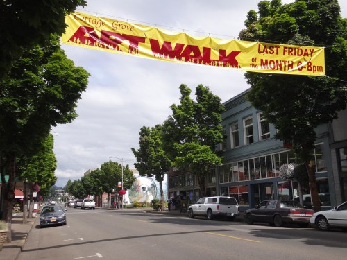A street view of Cottage Grove with a banner for the Art Walk event hanging above, lined with trees and parked cars.