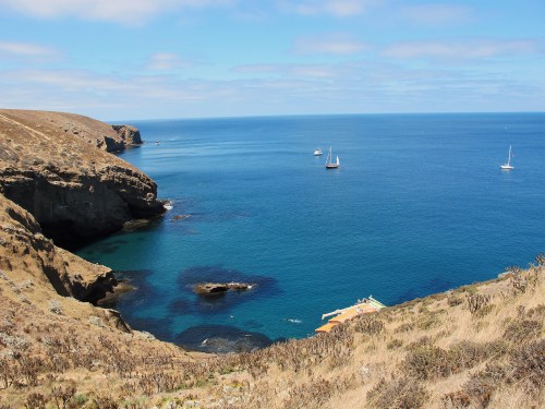 A serene coastal view with rocky cliffs, calm blue waters, and sailboats in the distance under a clear sky.