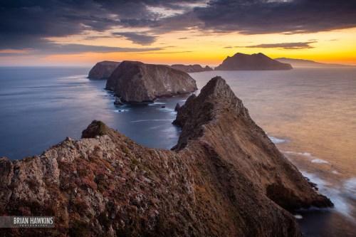 A dramatic coastal landscape at sunset, featuring rugged cliffs and distant islands under a colorful sky.