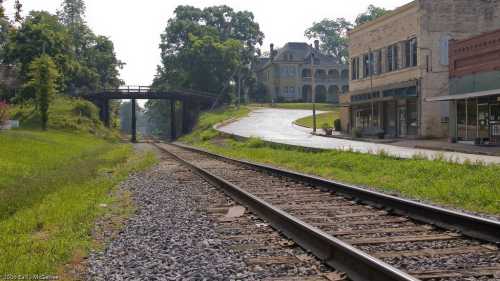 A view of a railway track beside a grassy area, with a bridge and a small town building in the background.