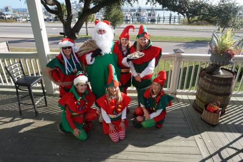 A group of festive elves and Santa pose together on a porch, with a marina visible in the background.