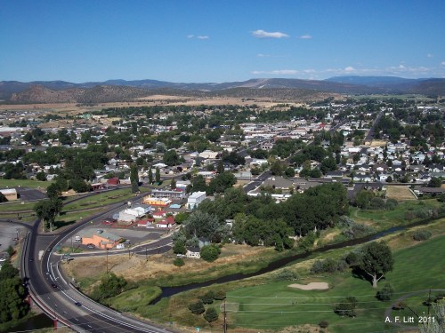 Aerial view of a small town surrounded by greenery and mountains, with roads and a river visible in the foreground.