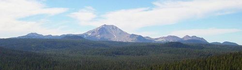 A panoramic view of a mountain range with a prominent peak, surrounded by dense green forests and a clear blue sky.