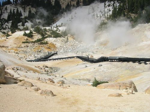 A wooden boardwalk winds through a rocky, steam-filled landscape with trees in the background.