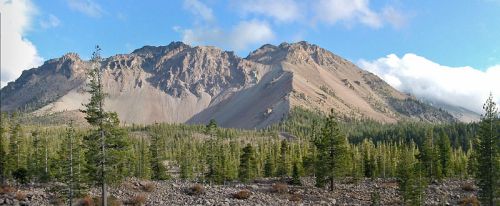 A panoramic view of a rugged mountain range surrounded by lush green trees under a clear blue sky.