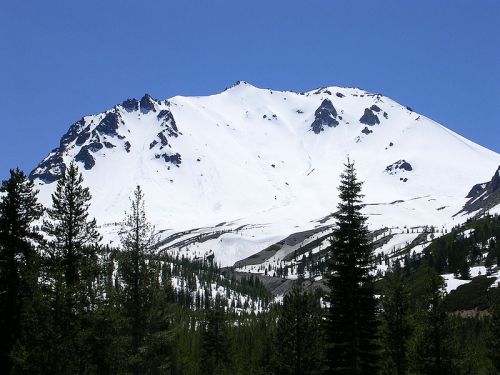 Snow-capped mountain under a clear blue sky, surrounded by evergreen trees and rugged terrain.