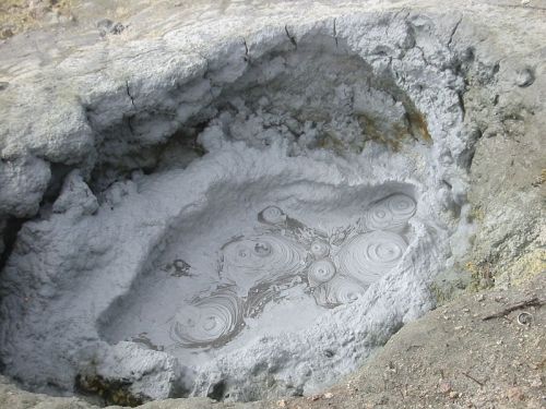 A shallow, gray mud pool with circular ripples, surrounded by rocky terrain.